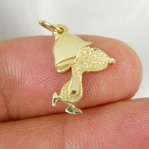 May include: A gold-colored charm in the shape of a cartoon character, with a textured surface. The charm features a loop at the top for attachment. The character is depicted in a walking pose, with a hat.