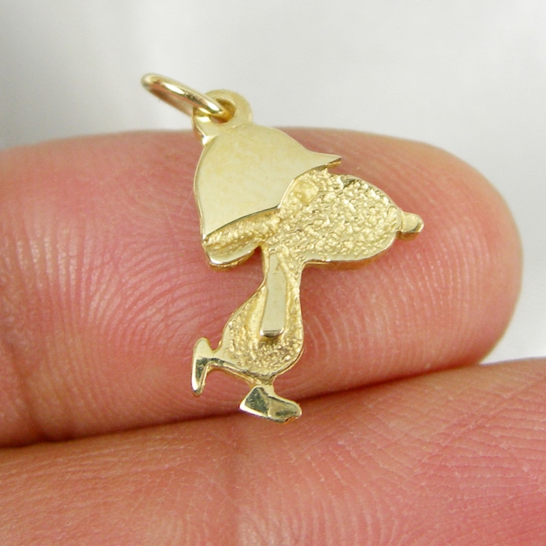 May include: A gold-colored charm in the shape of a cartoon character, with a textured surface. The charm features a loop at the top for attachment. The character is depicted in a walking pose, with a hat.