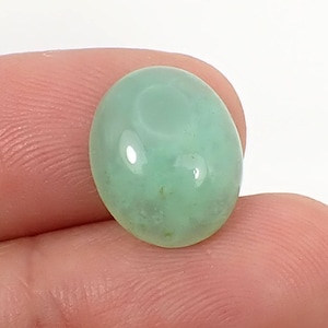 May include: A smooth, oval-shaped green gemstone with a light green hue and a white centre. The gemstone is polished and has a glossy finish.