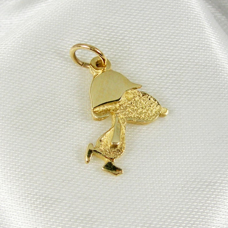 May include: A gold-colored charm in the shape of a cartoon character, with a textured body and a smooth hat. The charm is attached to a small loop, suggesting it's designed to be worn as a pendant or attached to a bracelet. The charm is set against a white background.