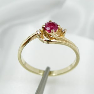 May include: A gold ring with a red ruby gemstone and two smaller diamonds. The ring has a delicate, twisted band.