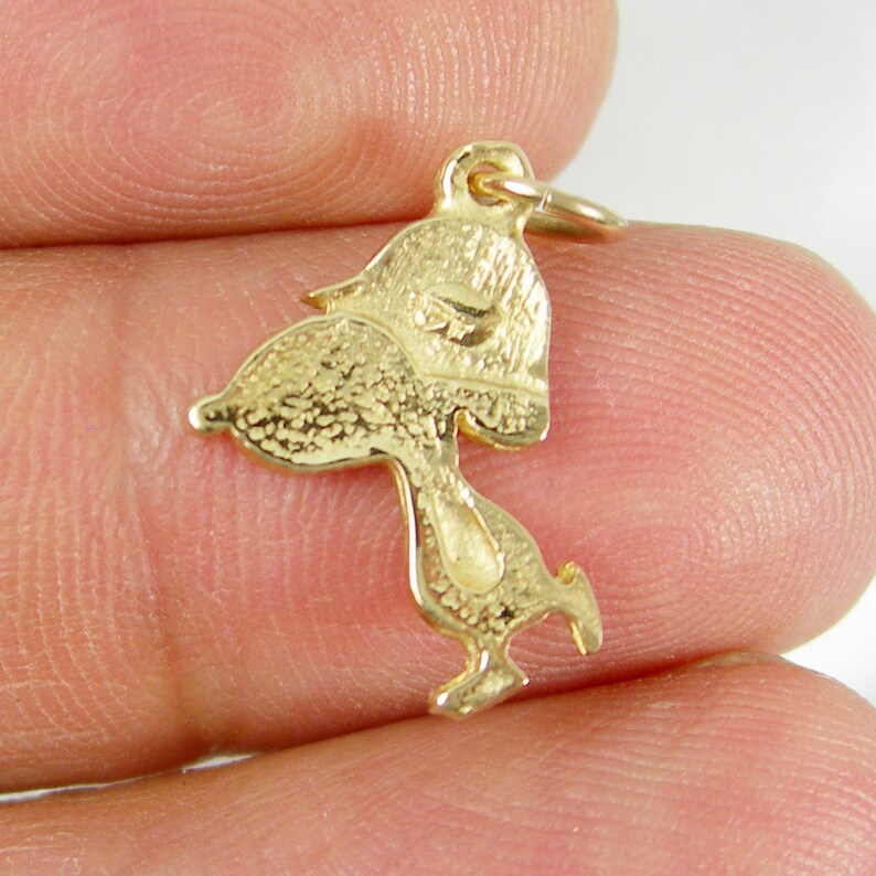 May include: A gold-colored charm in the shape of a cartoon character, possibly a bird. The charm has a textured surface and a small loop at the top for attachment to a necklace or bracelet. The charm is held in a person's hand.