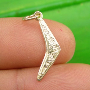May include: A small, gold-coloured boomerang charm with etched geometric patterns. The charm has a small loop at the top for attaching to a necklace or bracelet. The boomerang is held between two fingers.