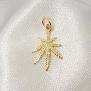 May include: A gold-colored pendant charm in the shape of a marijuana leaf. The leaf has a detailed design with veins and a textured surface.