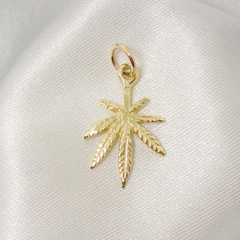 May include: A gold-colored pendant charm in the shape of a marijuana leaf. The leaf has a detailed design with veins and a textured surface.