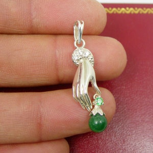 May include: A silver pendant featuring a hand holding a green jade sphere with a smaller green gemstone on top. The hand is adorned with small white gemstones.
