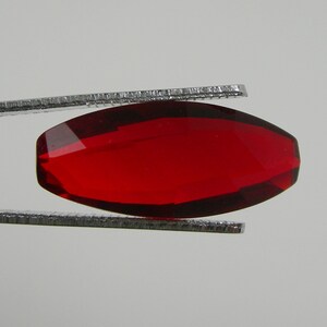 Puede incluir: Una gema facetada, alargada, de forma ovalada y color rojo intenso, es sostenida por unas pinzas plateadas. La gema tiene una superficie pulida y brillante, pareciendo transparente. Las facetas capturan la luz. Mide unos 2 cm.
