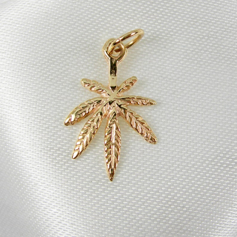 May include: A gold-colored pendant in the shape of a marijuana leaf. The leaf is detailed with veins and has a loop at the top for attaching to a chain.
