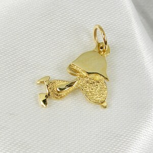 May include: A gold-colored pendant in the shape of a bellhop. The pendant has a textured body and a smooth, polished hat. The bellhop is depicted in a running pose, with legs extended. The pendant includes a small loop for attaching to a chain.