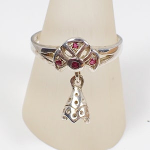 Peut inclure: Une bague en argent avec un pendentif. La bague présente un motif en forme d'éventail avec des pierres précieuses rouges et un petit pendentif en forme de cloche.