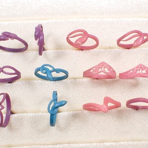 May include: A collection of various ring designs in shades of purple, blue, and pink. The rings feature diverse shapes, including knot, crown, and abstract designs. The rings are displayed on a white, textured surface.