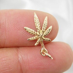 May include: A gold-colored marijuana leaf charm with a detailed design. The charm has a loop at the bottom for attaching to a necklace or bracelet.