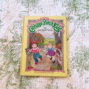 May include: A hardcover book titled Cabbage Patch Kids: The Just-Right Family. The yellow cover features an illustration of two children running through a forest, with the text A Parker Brothers Story Book, Story by Larry Callen, Pictures by Judy Gallen.