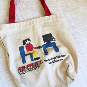 May include: A cream-colored canvas tote bag with red fabric handles. The front features a graphic of a person sitting at a desk with a computer, along with the text RE-PRINT LLC NATION'S LEADING DISCOUNTER 1-800-248-9171 Technology Solutions For Education.