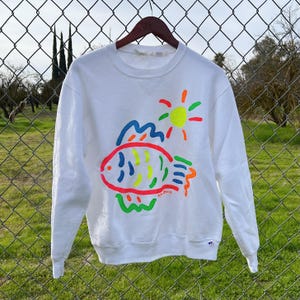 May include: White sweatshirt with a colorful graphic of a fish, sun, and seaweed. The graphic is in neon colors, including red, yellow, green, blue, and orange. The sweatshirt has a crew neck and long sleeves.