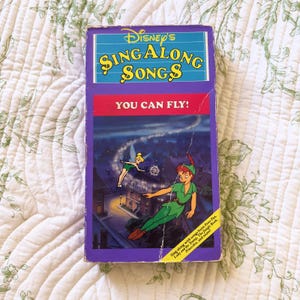 May include: A Disney Sing Along Songs VHS tape titled You Can Fly! featuring an illustration of Peter Pan and Tinker Bell flying over a night cityscape. The purple and red cover includes text listing songs from Peter Pan, Lady and the Tramp, and more.
