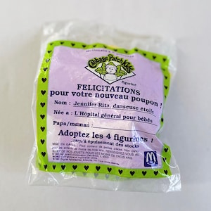 May include: Sealed Cabbage Patch Kids figurine in a clear plastic bag. The packaging is purple with green trim and features text in French, including "FELICITATIONS" and "Jennifer Rita, danseuse étoile." A McDonald's logo is visible.