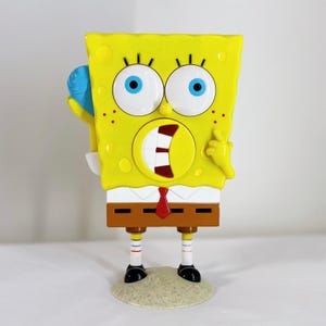 May include: A yellow SpongeBob SquarePants figurine with large blue eyes, a wide open mouth, and a red tie. The figurine is standing on a sandy base and holding a blue object. The figure has black shoes and brown pants.