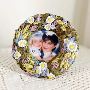 May include: Round photo frame with a floral wreath design. The frame features a picture of a woman and a child. The wreath is decorated with white daisies, blue and purple flowers, and green leaves. The frame is on a white surface.