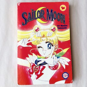 May include: A red Sailor Moon manga volume with the title in blue and yellow. The cover features a blonde anime character with blue eyes, making a peace sign. The author's name is also visible.