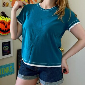 May include: A teal short-sleeved t-shirt featuring a white crew neckline, white sleeve cuffs, and a white hemline. The shirt has visible white contrast stitching along the neckline, sleeves, and bottom hem, creating a layered appearance.