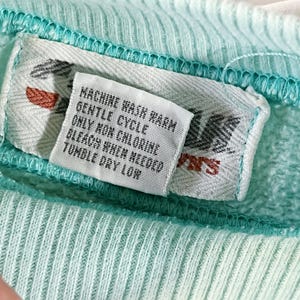 May include: A white care label with the text "Machine Wash Warm Gentle Cycle Only Non Chlorine Bleach When Needed Tumble Dry Low" on a light blue fabric.