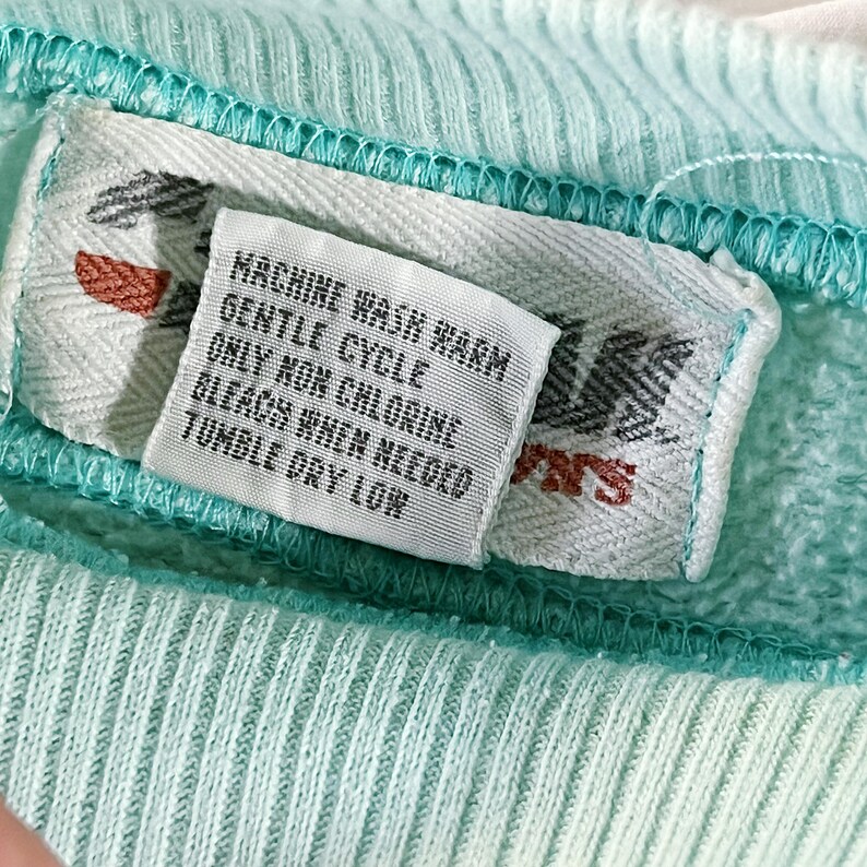 May include: A white care label with the text "Machine Wash Warm Gentle Cycle Only Non Chlorine Bleach When Needed Tumble Dry Low" on a light blue fabric.