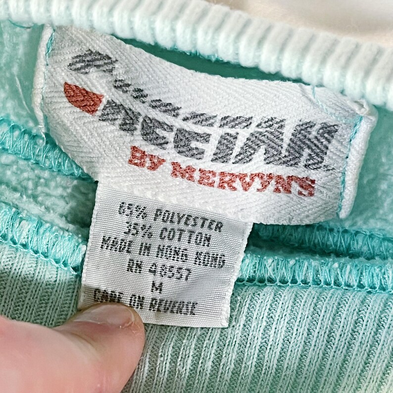 May include: A white tag with black text that reads "Ocean by Mervyns" and details the fabric composition as 65% polyester and 35% cotton. The tag also states "Made in Hong Kong" and "RN 43557". The tag is attached to a light blue sweatshirt.