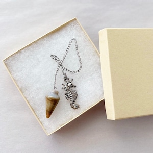 May include: A pendulum with a silver chain and seahorse charm rests in an open beige gift box. The pendulum has a cone-shaped, brown and white stone. The seahorse charm is detailed with a crown.
