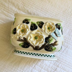 May include: A cream-colored ceramic planter featuring raised, three-dimensional white magnolia flowers with yellow centers, dark green leaves, and a light blue butterfly. The base is decorated with a green and white checkered border pattern.