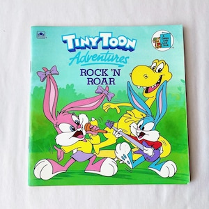 May include: A children's book titled "Tiny Toon Adventures Rock 'N Roar" features cartoon characters. The cover shows a pink bunny with a microphone, a blue bunny with a guitar, and a yellow dinosaur. The background is green and blue.