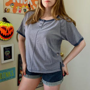 May include: A short-sleeved henley shirt featuring a navy blue and white horizontal striped pattern, a round neckline with three buttons, and solid navy blue trim on the collar and sleeve cuffs, paired with dark blue denim cuffed shorts.