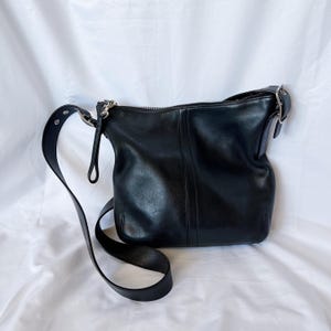 May include: Black leather shoulder bag with a long adjustable strap. The bag has a zipper closure and silver-tone hardware. The bag is on a white background.