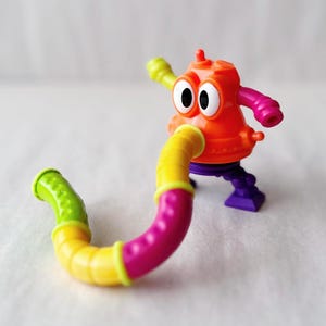 May include: A colorful plastic toy robot with a flexible, segmented tube. The robot has an orange body, large white eyes, and arms in green and pink. The tube is yellow, pink, and green. The toy stands on purple feet.