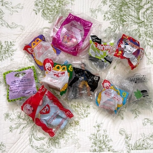 May include: A collection of ten assorted McDonald's Happy Meal toys, each individually sealed in clear plastic packaging. The set includes various branded items featuring characters from Disney, Hot Wheels, Peanuts, and Ty, with visible text.