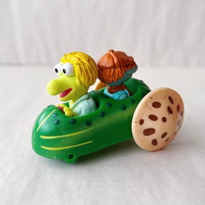 May include: A green toy car with yellow trim and a single wheel. Two cartoon characters are seated inside. One character has yellow hair and a long nose, and the other has red hair and a brown hat. The wheel is tan with brown spots.