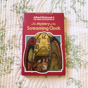 May include: A paperback book titled Alfred Hitchcock and The Three Investigators in The Mystery of the Screaming Clock. The red cover features a large yellow clock illustration containing three figures and smaller clock shapes in the background.