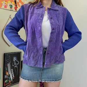 May include: A purple suede jacket featuring a collared neckline, a button-down front, and long blue ribbed knit sleeves. The jacket has a textured suede body with visible stitching details along the shoulders and chest, paired with a white shirt.