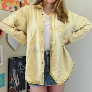 May include: A long-sleeved button-down shirt in pale yellow featuring a white undershirt, a denim mini skirt, and decorative yellow patterned trim along the front placket and hem. The shirt includes light-colored embroidered geometric accents.