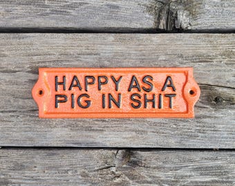 May include: Orange rectangular sign with the phrase "HAPPY AS A PIG IN SHIT" in black lettering. The sign has two holes on each end for mounting. The sign is set against a weathered wood background.