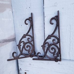Puede incluir: Dos soportes de estante de hierro fundido adornados con un dise&ntilde;o de desplazamiento. Los soportes son de color marr&oacute;n oscuro y tienen un acabado r&uacute;stico.