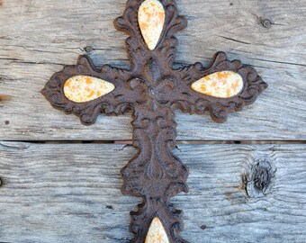 May include: A brown cast iron cross with four white and orange teardrop-shaped accents. The cross is hanging on a wooden wall.