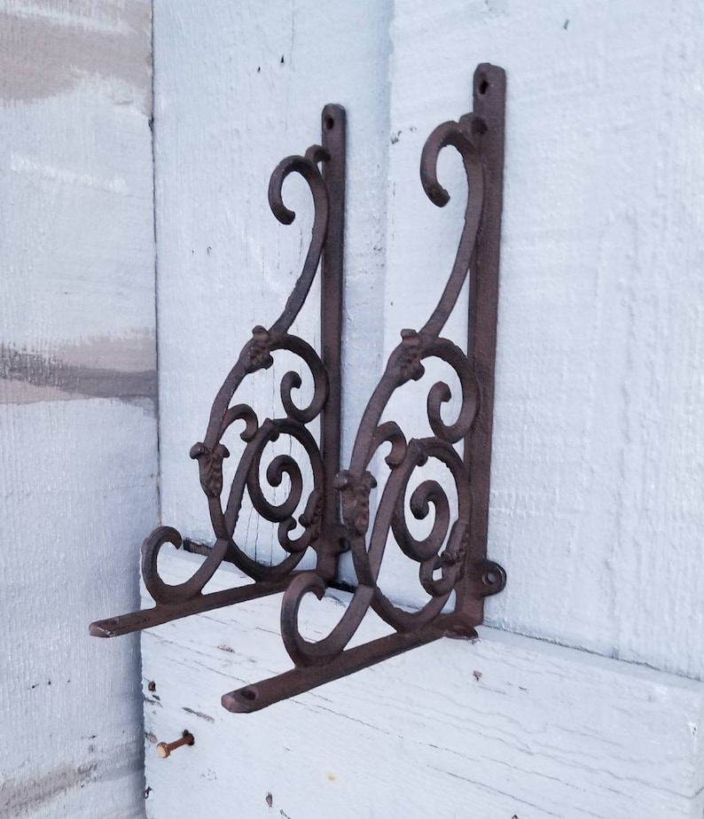 Puede incluir: Dos soportes de estante de hierro fundido ornamentados con un dise&ntilde;o de desplazamiento. Los soportes son de color marr&oacute;n oscuro y tienen un acabado r&uacute;stico.