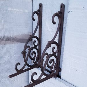 Puede incluir: Dos soportes de estante de metal adornados con un dise&ntilde;o de desplazamiento. Los soportes son de color marr&oacute;n oscuro y est&aacute;n montados en una pared de madera blanca.