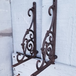 Puede incluir: Dos soportes de estante de hierro fundido adornados con un dise&ntilde;o de desplazamiento. Los soportes son de color marr&oacute;n oscuro y tienen un acabado r&uacute;stico.
