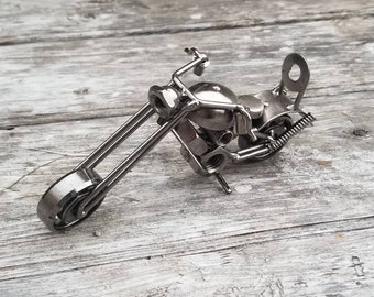 May include: A silver metal motorcycle sculpture made from nuts, bolts, and other hardware. The motorcycle is on a weathered wooden surface.