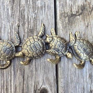 May include: A decorative bronze-colored metal wall hanging featuring four stylized sea turtles. Each turtle has a textured shell and is arranged in a horizontal line. The turtles are mounted on a weathered wooden surface.