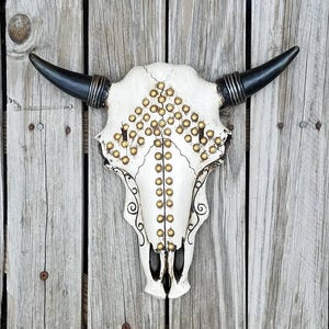 Peut inclure: Cr&acirc;ne de taureau blanc avec des clous dor&eacute;s et des accents noirs, mont&eacute; sur un mur en bois.