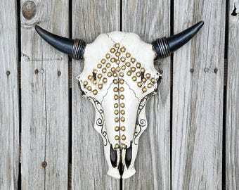 May include: White bull skull with gold studs and black accents, mounted on a wooden wall.