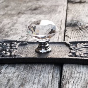 May include: A decorative cabinet pull with a clear crystal knob and a black, ornate backplate. The backplate features floral designs on each end. The hardware is designed for furniture and home decor.